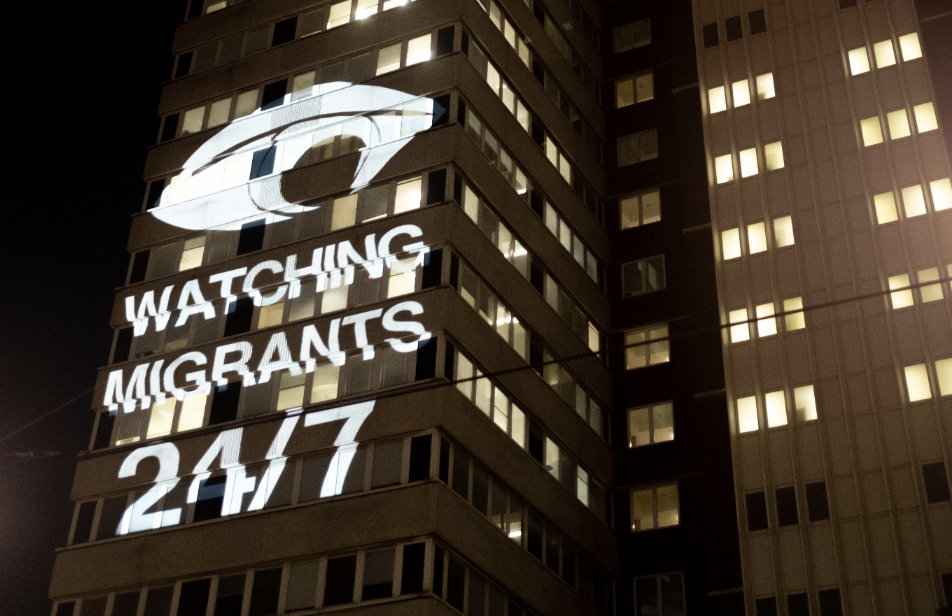Photo of Lunar House (immigration centre) with light projection of text reading: Stop Watching Migrants 24/7