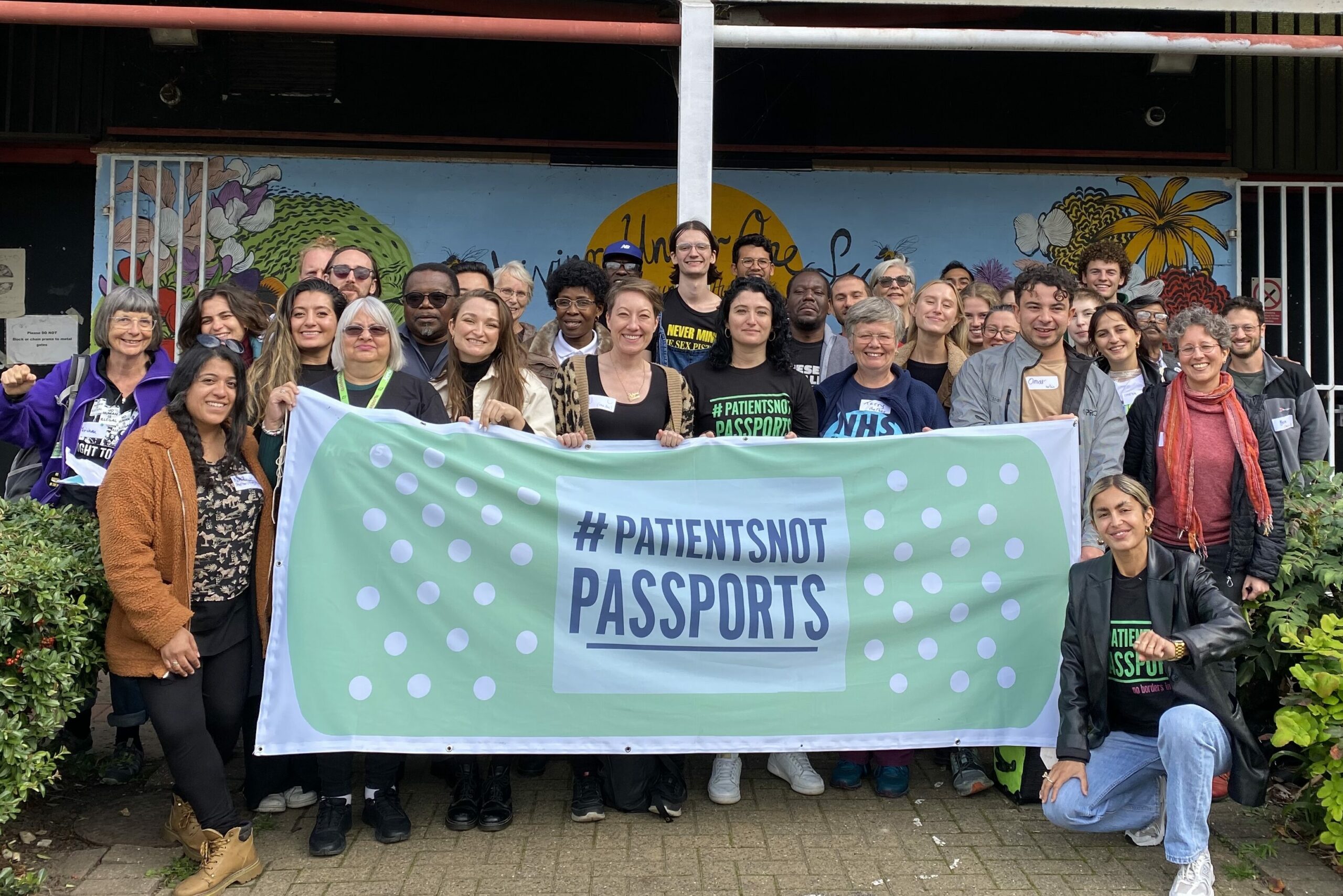 Image of Patients Not Passports campaigners with a green banner saying 'Patients Not Passports' outside Living Under One Sun Community Centre
