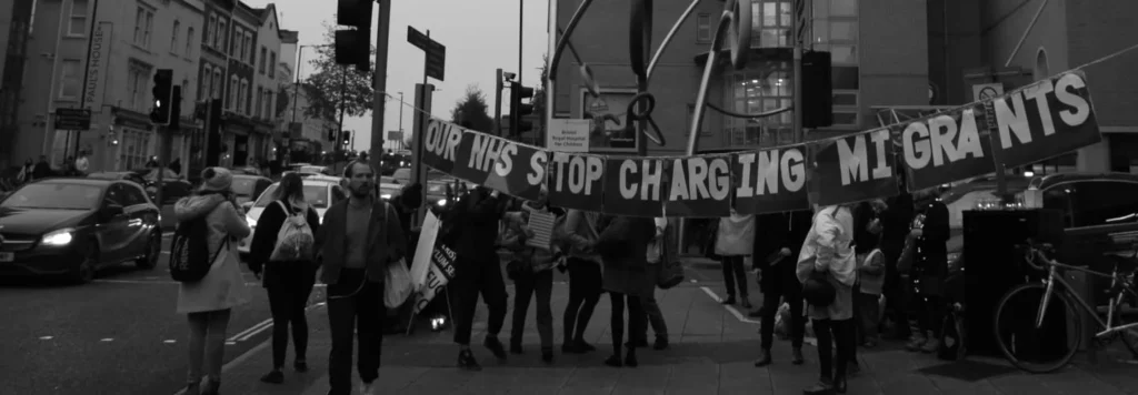 A black and white photograph of people on a street with a banner that says Our NHS Stop Charging Migrants